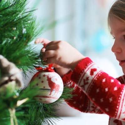 A young child wearing a red and white sweater hangs an ornament on a Christmas tree