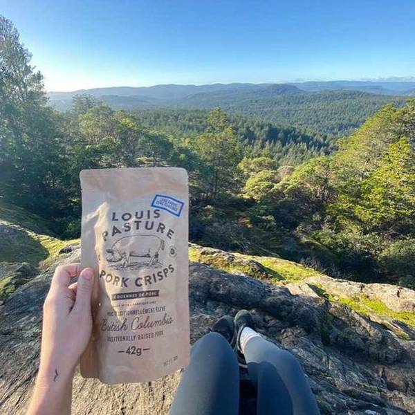 A person sitting on a mountain holds a bag of Louis Pasture Pork Crisps in Vancouver Island.