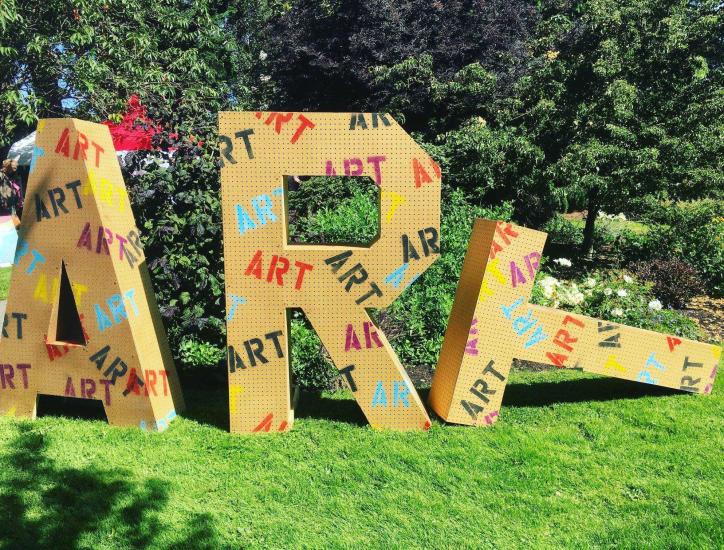 Three large, yellow, block letters spell out "ART" with the "T" toppled over on the green grass.