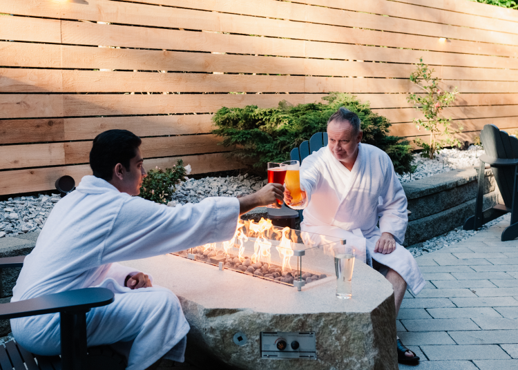 Two men sit on a patio in white bathrobes around a gas fire and are clinking beers together in a toast.