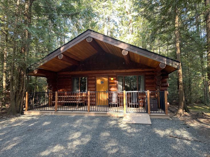 An exterior view of a log cabin cottage surrounded by evergreens in the sunshine at Tigh-Na-Mara Seaside Spa and Resort.