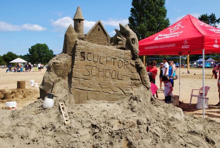 "Sculptor School" is carved into the front of an expert sand castle with tools strewn about and a red shade tent nearby.