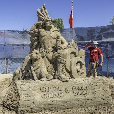 Huge sand castle of Captain Canada & Beaver Buddy, with a man next to it raising his arm to signify strength.