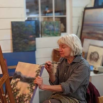 a woman sits in an art studio, painting the edge of a canvas depicting an outdoor scene at Tosh Art Centre