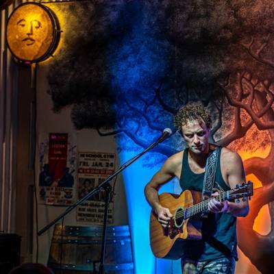 A musician, Adrian Chalifour, plays acoustic guitar on an indoor stage with blue and orange backlights