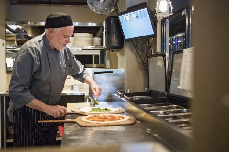 Eric Edwards, a chef wearing a gray uniform, creates two different pizzas in the kitchen at Tigh Na Mara