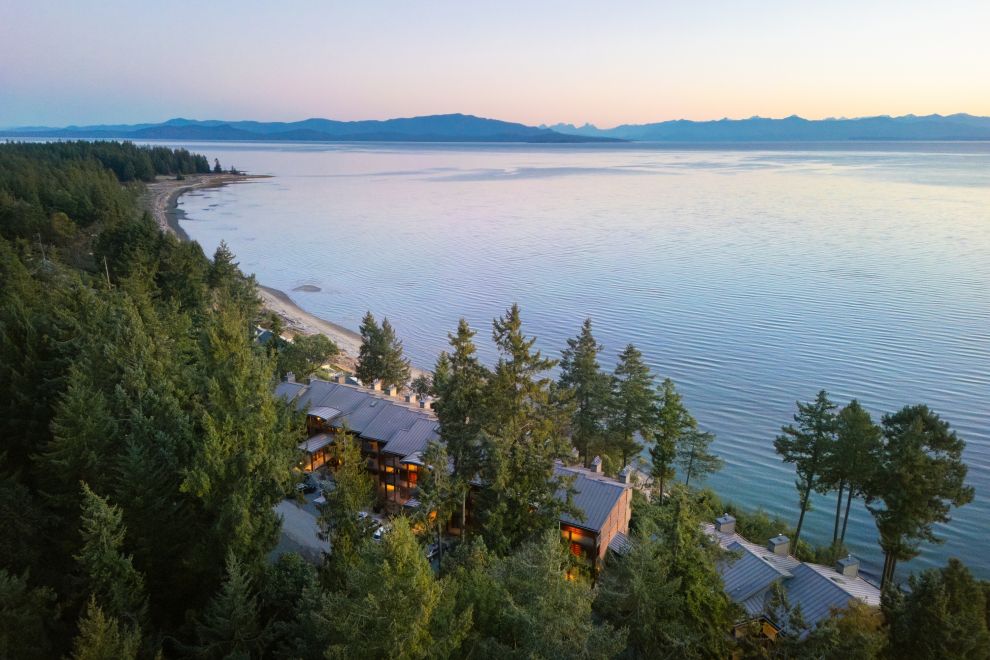 An aerial view above Tigh-Na-Mara Seaside Spa Resort with tall evergreens beside the calm open ocean at dusk.