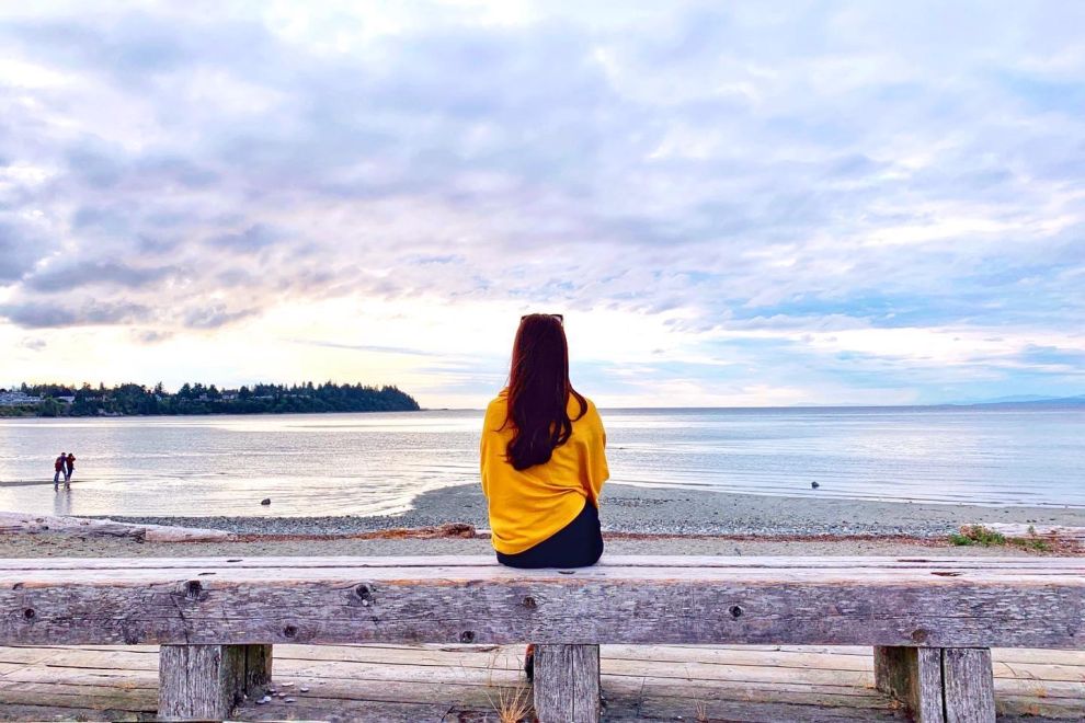 a woman in a yellow jacket sits on a bench and looks out at the beach in Parksville Qualicum Beach