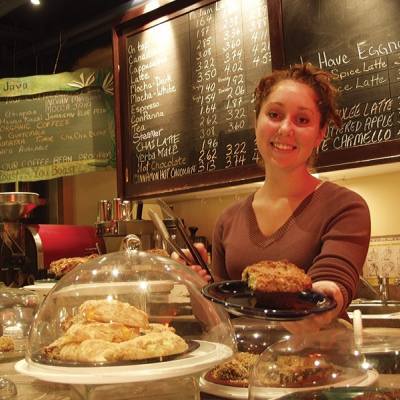 Woman hands a pastry to the camera while working at a coffee bar. There are several dessert stands filled with pastries