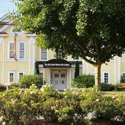 bushes and a tree stand in front of the yellow exterior of the TOSH - the old school house in qualicum beach